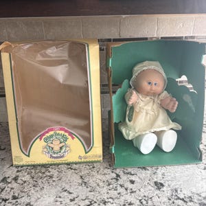 Op de afbeelding: Een vintage Cabbage Patch Kids Preemie pop uit 1985 in de originele doos. De pop heeft blauwe ogen, een crèmekleurige jurk en een bijpassend mutsje. De doos is geel met groene accenten en het Cabbage Patch Kids logo.