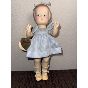 May include: A vintage Patsy doll in a blue and white gingham dress, with a matching bow and cream-coloured shoes. The doll has a heart-shaped tag that reads "Patsy". The doll is standing on a brown surface.