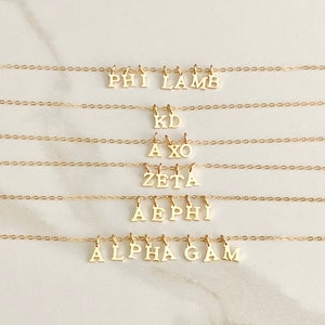 May include: Five gold necklaces with different Greek letters hanging from them. The necklaces are arranged in a vertical stack, with the top necklace being the shortest and the bottom necklace being the longest. The letters on the necklaces are: Phi Lamb, KD, AX, Zeta, AEPhi, and Alpha Gam.