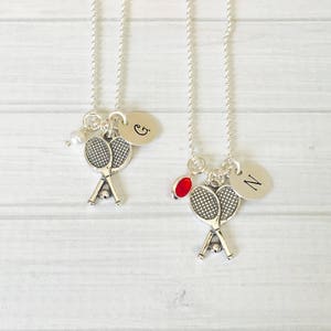 May include: Two silver tennis racket charm necklaces. One necklace has a pearl and a charm with the letter "G". The other necklace has a red charm and a charm with the letter "N". Both necklaces have a ball chain.