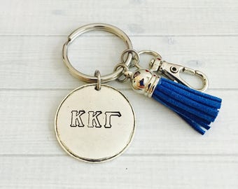 May include: Silver keychain with a round charm engraved with the letters "KKГ" and a blue tassel.