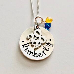 May include: A silver necklace with a round pendant featuring the year "2020" and the name "Kimberly" in cursive. The necklace has a delicate chain and is adorned with yellow and blue crystal beads.