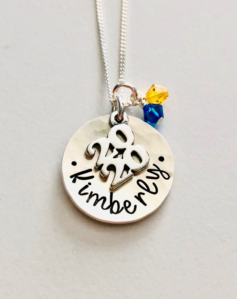 May include: A silver necklace with a round pendant featuring the year "2020" and the name "Kimberly" in cursive. The necklace has a delicate chain and is adorned with yellow and blue crystal beads.