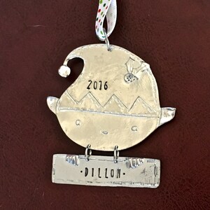 May include: Silver elf ornament with a stitched hat, a bell, and the year 2016 engraved on the front. A separate silver tag hangs below with the name "DILLON" engraved on it.