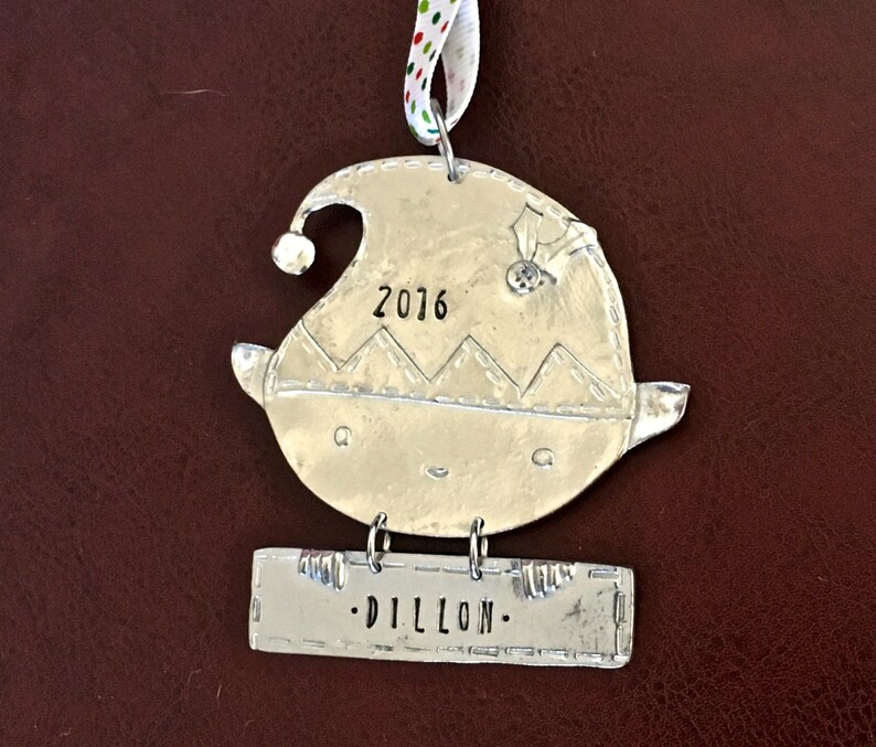 May include: Silver elf ornament with a stitched hat, a bell, and the year 2016 engraved on the front. A separate silver tag hangs below with the name "DILLON" engraved on it.