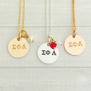 May include: Three gold, silver, and rose gold necklaces with round pendants that say "ΣΦΛ". The gold necklace has a small pearl charm, the silver necklace has a red crystal charm, and the rose gold necklace has a small charm.