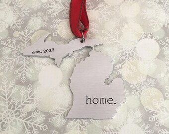 May include: A silver metal ornament shaped like the state of Michigan with the text "est. 2017" and "home." on it. The ornament is hanging from a red ribbon with white stitching.