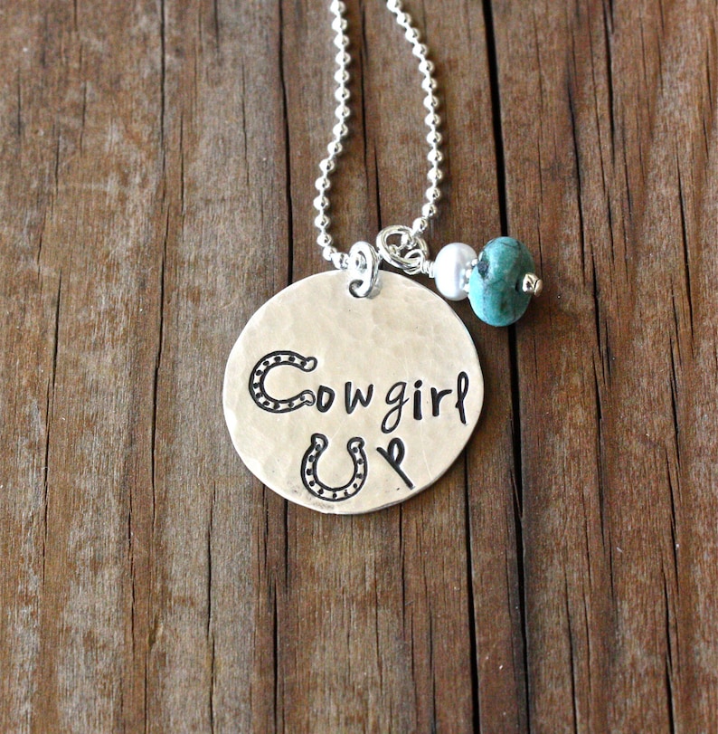 May include: Silver-toned pendant necklace with the words "Cowgirl Up" and a horseshoe design. The pendant is round and has a hammered texture. The necklace includes a turquoise bead and a white bead.