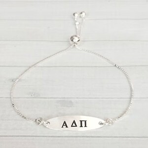 May include: A silver adjustable bracelet with a silver oval charm that says "AΔΠ".