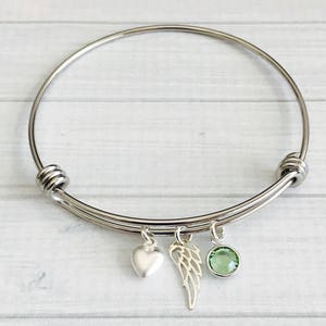 May include: A silver bangle bracelet with three charms: a heart, a wing, and a green crystal.