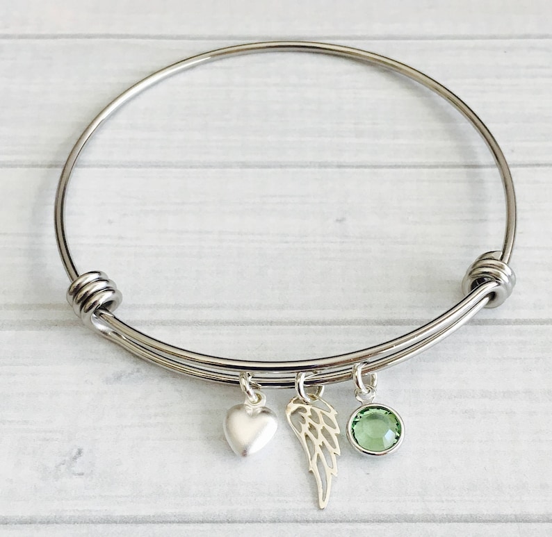 May include: A silver bangle bracelet with three charms: a heart, a wing, and a green crystal.