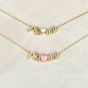 May include: Two gold-colored necklaces with the letters "phi mu" in a cursive font. The heart-shaped charm is either white or pink. The necklaces are on a light-colored surface.