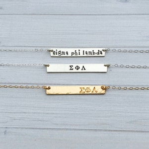 May include: Three necklaces with rectangular pendants. The top necklace has a silver chain and pendant with the text "sigma phi lambda". The middle necklace has a silver chain and pendant with the Greek letters "ΣΦΛ". The bottom necklace has a gold chain and pendant with the Greek letters "ΣΦΛ".