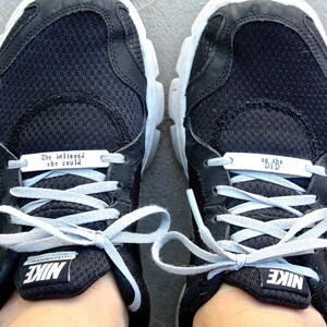 May include: A pair of black Nike sneakers with white laces. The left shoe has a silver tag that reads "She believed she could" and the right shoe has a silver tag that reads "So she did."