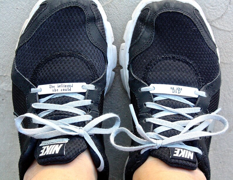 May include: A pair of black Nike sneakers with white laces. The left shoe has a silver tag that reads "She believed she could" and the right shoe has a silver tag that reads "So she did."