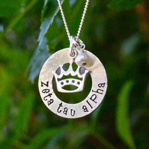 May include: Silver necklace with a round pendant that says "Zeta Tau Alpha" and has a crown cut out in the center. A small pearl charm hangs from the pendant.