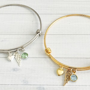 May include: Two delicate bangle bracelets with charms. One bracelet is silver with a green crystal and a wing charm. The other bracelet is gold with a blue crystal and a wing charm.