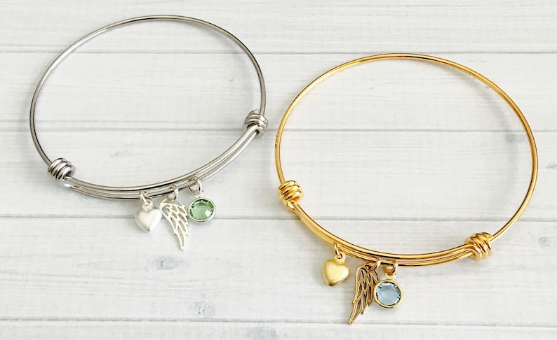May include: Two delicate bangle bracelets with charms. One bracelet is silver with a green crystal and a wing charm. The other bracelet is gold with a blue crystal and a wing charm.