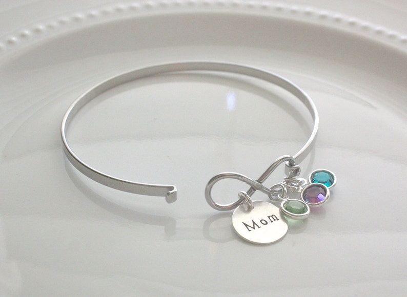 Może przedstawiać: Srebrna bransoletka typu bangle z zawieszką w kształcie nieskończoności i małą okrągłą zawieszką z napisem "Mom". Bransoletka posiada kilka kolorowych zawieszek z kamieni szlachetnych.