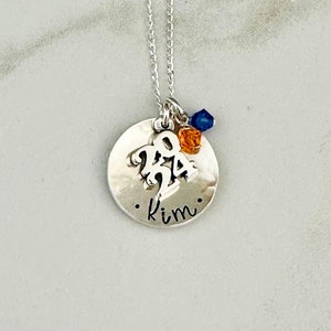 May include: A silver necklace with a round pendant. The pendant has the number "2024" cut out, the name "Kim" engraved, and is adorned with blue and orange beads. The necklace chain is delicate.