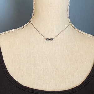 Peut inclure: Un collier en argent avec un pendentif en forme d'halt&egrave;re. Le collier est port&eacute; par un mannequin portant un d&eacute;bardeur noir.