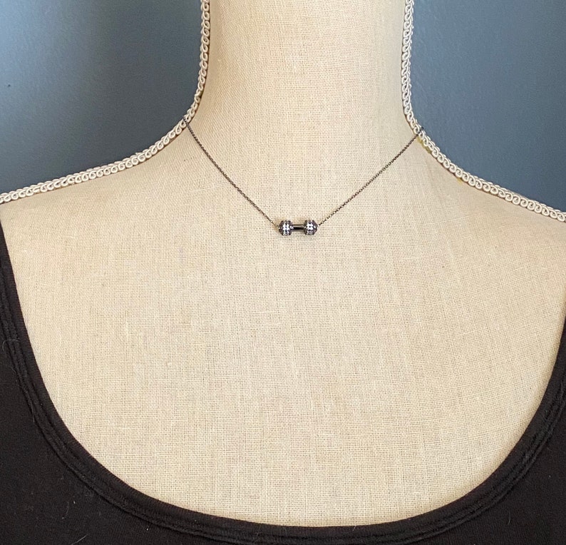 Peut inclure: Un collier en argent avec un pendentif en forme d'halt&egrave;re. Le collier est port&eacute; par un mannequin portant un d&eacute;bardeur noir.