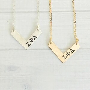 May include: Two silver and gold necklaces with a chevron pendant. The pendant is engraved with the Greek letters Sigma Phi Lambda.