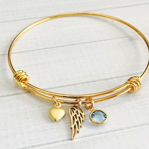 May include: A gold expandable bangle bracelet with a heart, angel wing, and light blue crystal charm.