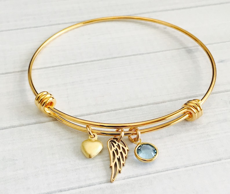 May include: A gold expandable bangle bracelet with a heart, angel wing, and light blue crystal charm.