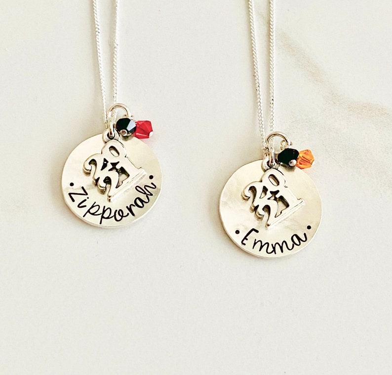 May include: Two silver necklaces with round pendants. Each pendant has a '22' charm, a name, and a small bead cluster. One necklace says 'Zipporah', the other says 'Emma'. The chains are delicate.