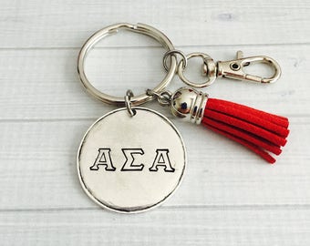 May include: Silver-toned keychain with a round charm featuring the letters AΣA. A bright red tassel hangs from the keychain, which also includes a clasp and a key ring. The keychain is a decorative accessory.