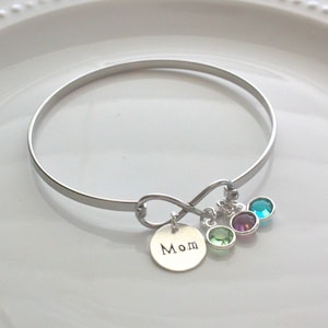 Może przedstawiać: Srebrna bransoletka typu bangle z zawieszką w kształcie nieskończoności i małą okrągłą zawieszką z napisem "Mom". Bransoletka posiada trzy kolorowe zawieszki z kamieni szlachetnych w kolorze zielonym, fioletowym i turkusowym. Prosta i elegancka biżuteria.