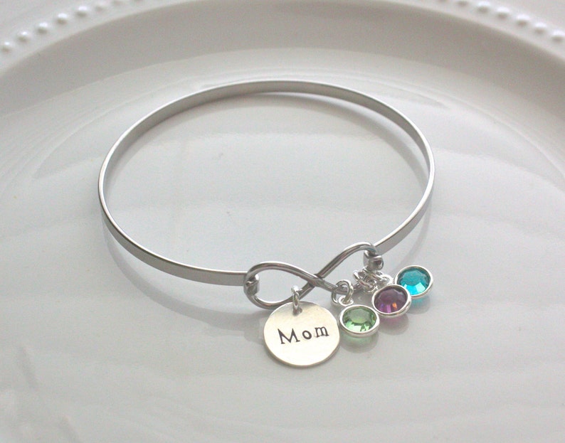 Może przedstawiać: Srebrna bransoletka typu bangle z zawieszką w kształcie nieskończoności i małą okrągłą zawieszką z napisem "Mom". Bransoletka posiada trzy kolorowe zawieszki z kamieni szlachetnych w kolorze zielonym, fioletowym i turkusowym. Prosta i elegancka biżuteria.