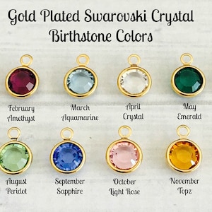 May include: Gold plated Swarovski crystal birthstone charms in various colors, arranged in rows by month. Each charm features a round crystal with a gold setting. The charms are displayed on a light wood background.