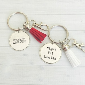 May include: Two silver keychains with round pendants. One keychain has a red tassel, the other a white tassel. The pendants have stamped text: "ΣΦΛ" and "Sigma Phi Lambda". Each keychain has a clasp.
