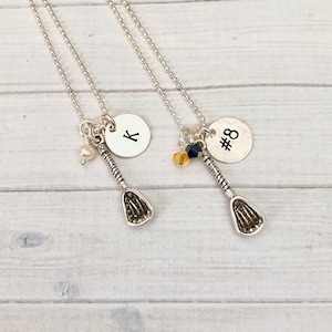 May include: Two silver necklaces with lacrosse stick charms. One necklace has a round charm with the letter "K" and a pearl. The other has a round charm with the number "#8" and two small beads.