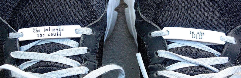 May include: A pair of black sneakers with white laces. The left shoe has a silver tag that reads "She believed she could". The right shoe has a silver tag that reads "So she did".