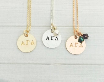 May include: Three gold necklaces with round pendants featuring the letters "AGD". The pendants are on different colored chains: gold, silver, and rose gold. The rose gold necklace has a green crystal charm.