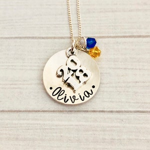 May include: A silver-colored necklace with a round pendant. The pendant features the name "Olivia" in cursive, a "2023" charm, and blue and yellow crystal accents. The necklace hangs on a delicate chain.