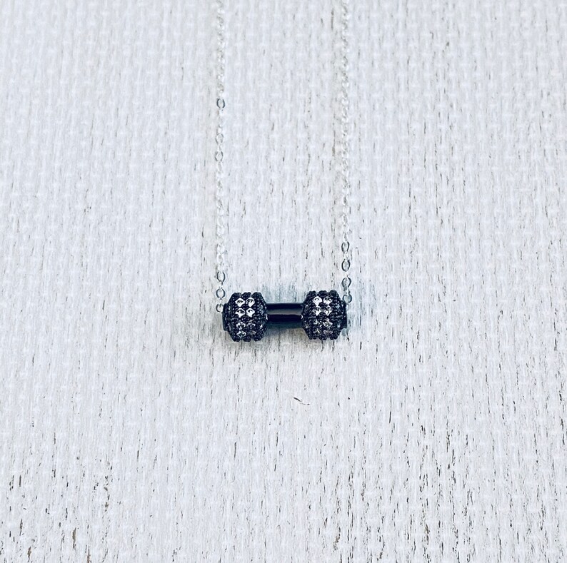 Peut inclure: Un collier en cha&icirc;ne argent&eacute;e avec un pendentif en forme d'halt&egrave;re noir. L'halt&egrave;re est recouvert de petits cristaux transparents.