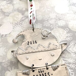 May include: Silver Christmas ornament shaped like an elf holding a sign that says "the . BUDDES.". The elf has a stitched hat with the year "2016" on it.