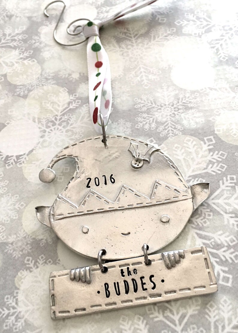 May include: Silver Christmas ornament shaped like an elf holding a sign that says "the . BUDDES.". The elf has a stitched hat with the year "2016" on it.