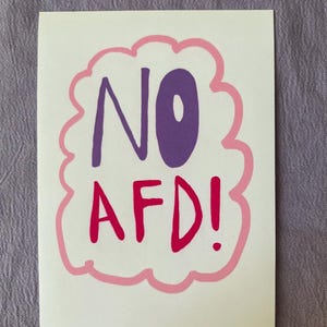 Könnte beinhalten: Ein weißer Aufkleber mit den Worten "NO AFD!" in Lila und Rot, innerhalb einer rosa und hellrosa wolkenförmigen Umrandung. Der Aufkleber befindet sich auf einer lila Oberfläche.
