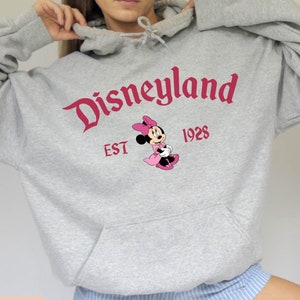 May include: A light grey Disneyland hoodie with a kangaroo pocket. The front features the Disneyland logo in red, the year 1928, and a Minnie Mouse graphic. The words "SPORT GREY" are printed at the bottom.