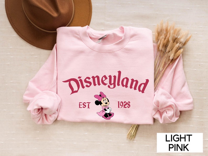 May include: Light pink Disneyland sweatshirt with the Disneyland logo in burgundy script, the year 1928, and a Minnie Mouse graphic. The sweatshirt is long-sleeved and has a crew neck. A brown hat and dried flowers are in the background.