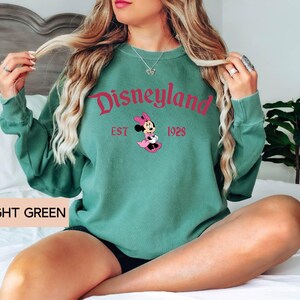 May include: Light green sweatshirt with the word "Disneyland" in pink script, along with "EST 1928" and a Minnie Mouse graphic. The text "LIGHT GREEN" is printed at the bottom. The sweatshirt has long sleeves.