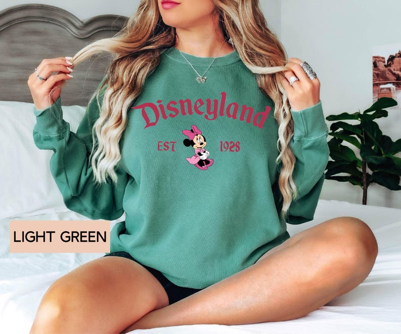 May include: Light green sweatshirt with the word "Disneyland" in pink script, along with "EST 1928" and a Minnie Mouse graphic. The text "LIGHT GREEN" is printed at the bottom. The sweatshirt has long sleeves.