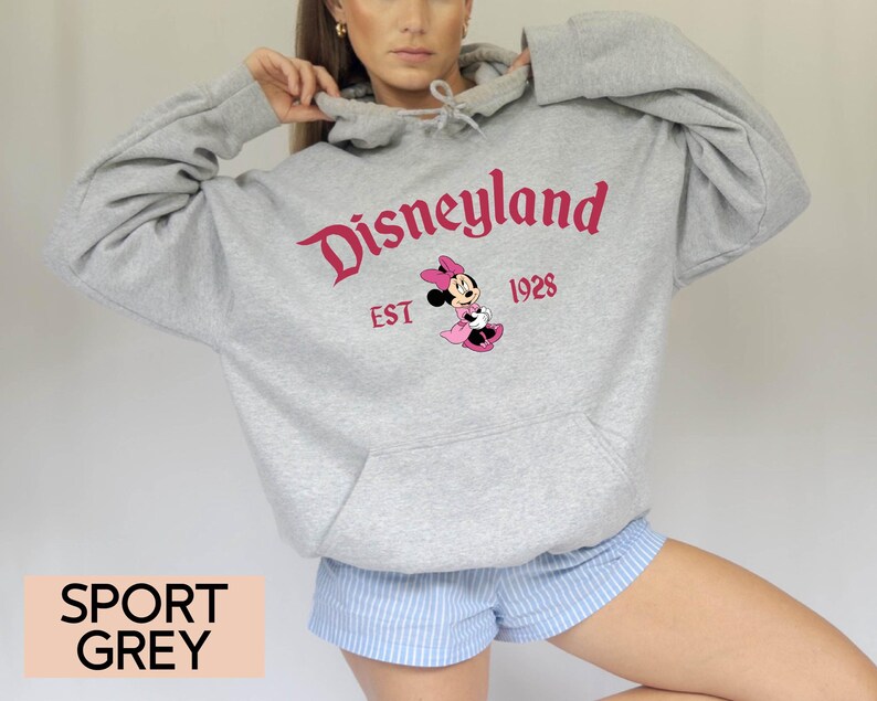 May include: A light grey Disneyland hoodie with a kangaroo pocket. The front features the Disneyland logo in red, the year 1928, and a Minnie Mouse graphic. The words "SPORT GREY" are printed at the bottom.