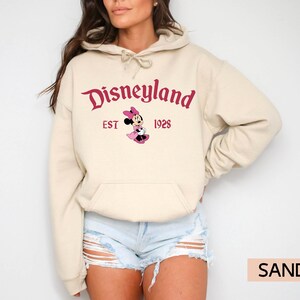 May include: Sand-colored hoodie featuring the Disneyland logo in burgundy script, with "EST 1928" below. A Minnie Mouse graphic is centered under the text. The hoodie has a drawstring hood and a front pocket.