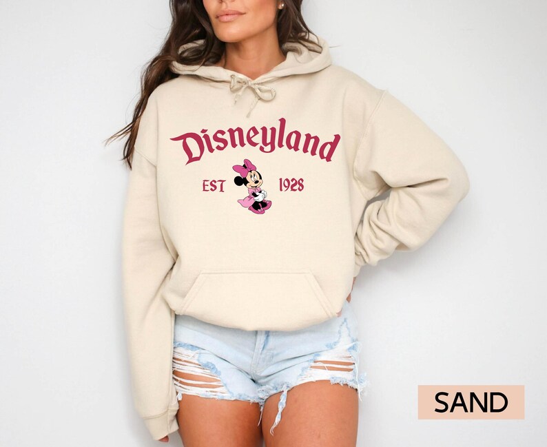 May include: Sand-colored hoodie featuring the Disneyland logo in burgundy script, with "EST 1928" below. A Minnie Mouse graphic is centered under the text. The hoodie has a drawstring hood and a front pocket.
